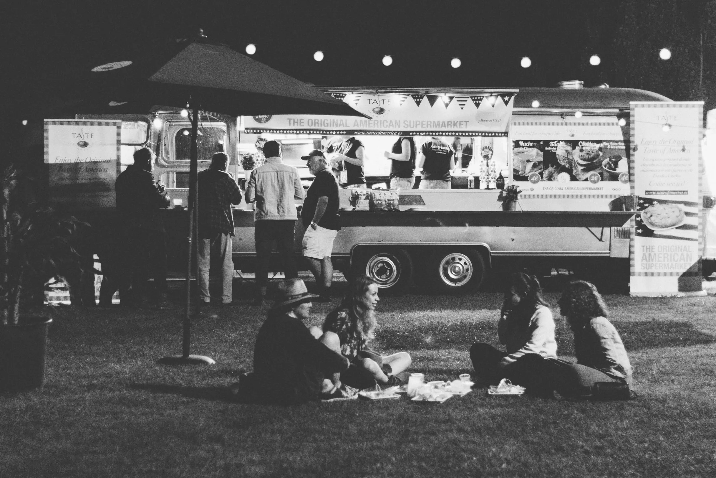 Un food truck éclairé de nuit, avec des personnes qui commandent et des convives assis sur l’herbe